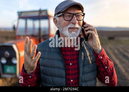 Excited mature farmer talking on mobile phone in front of tractor in field Stock Photo