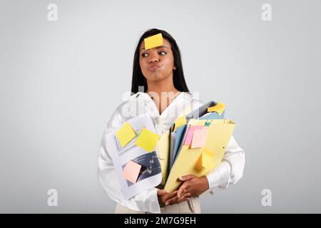 Tired busy millennial black female in white blouse hold many folders of documents, coffee cup, has sticker on forehead Stock Photo