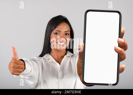 Smiling confident millennial black female in white blouse show thumb up and smartphone with empty screen Stock Photo