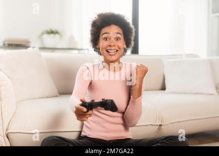 Cheerful excited young mixed race lady with joystick playing computer game, make victory and success gesture Stock Photo