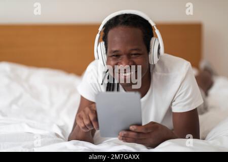 Happy Black Man With Digital Tablet And Wireless Headphones Relaxing In Bed Stock Photo