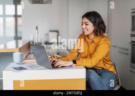 Remote Work. Smiling Young Arab Female Freelancer Working With Laptop In Kitchen Stock Photo