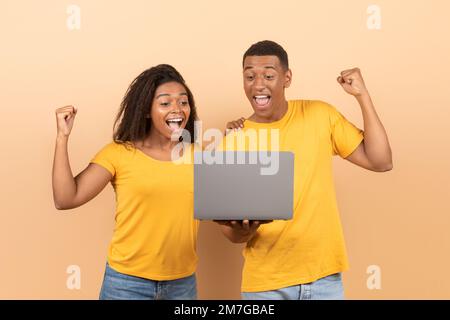 Yeah. Overjoyed black spouses celebrating online win, great deal, holding laptop and shaking clenched fists Stock Photo