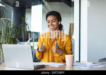 E-teaching. Happy black female business tutor gesturing thumbs up to laptop computer webcamera, sitting in office Stock Photo
