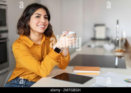 Smiling Young Arab Woman Drinking Coffee And Using Digital Tablet In Kitchen Stock Photo