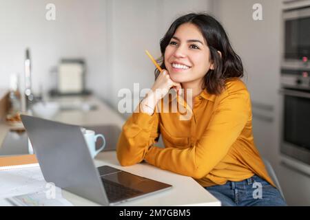 Dreamy Young Arab Female Sitting At Desk With Laptop In Kitchen Stock Photo