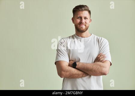 Waist up portrait of bearded young man smiling at camera while standing with arms crossed against pastel green, minimal Stock Photo
