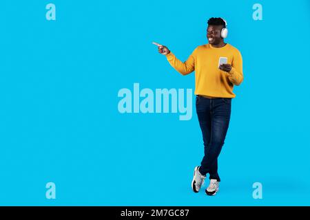 Black Man Wearing Headphones Holding Smartphone In Hand And Pointing Aside Stock Photo