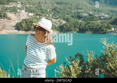 Smiling woman in hat and sunglasses with wild hair standing near mountains lake on background. Positive young woman traveling on blue lake outdoors Stock Photo