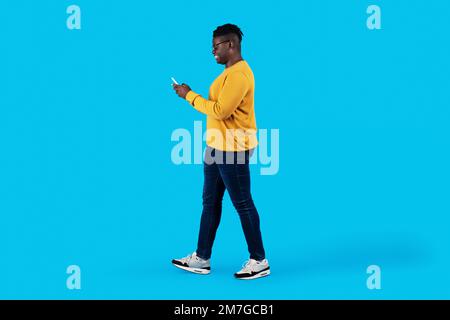 Young Black Man Walking With Smartphone In Hands Over Blue Studio Background Stock Photo