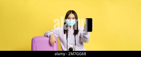 Image of girl traveller, asian tourist shows her phone screen, covid health passport on smartphone app, wearing medical mask and suitcase, yellow Stock Photo