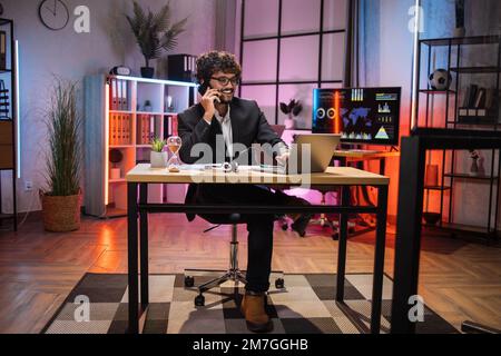 Working process at office of young serious guy having phone call with partners at evening time. Positive bearded businessman having mobile conversation while sitting at table and working on laptop. Stock Photo