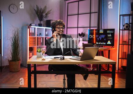 Working process at office of young serious guy having phone call with partners at evening time. Positive bearded businessman having mobile conversation while sitting at table and working on laptop. Stock Photo