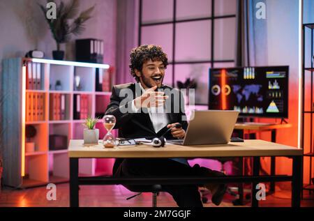 Positive excited young manager or businessman, having successful deal during video call with business partners, wearing suit sitting at table, using laptop at evening time in modern office. Stock Photo
