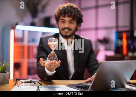 Smiling confident businessman in formal suit sitting at desk with modern laptop and holding hourglass in his hand. Concept of deadline people and office work. Stock Photo