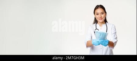 Portrait of asian female doctor, nurse in rubber gloves, giving sterile medical face masks and smiling, measures to prevent coronavirus disease, white Stock Photo