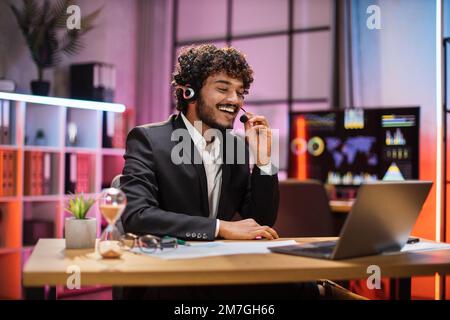 Positive man in headset having video call with partners at evening time, working process at office. Positive bearded businessman having conversation while sitting at table with laptop. Stock Photo