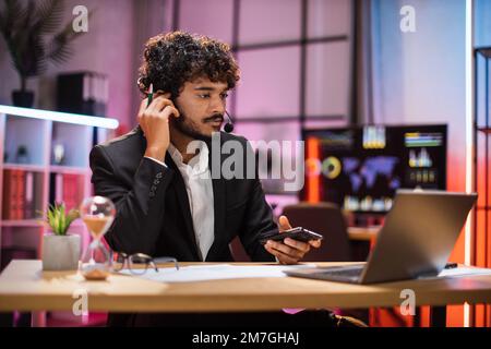 Deadline concept of project, working at evening time in office. Serious businessman, broker, financial expert in suit and headset working using laptop computer. Stock Photo