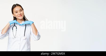 Image of smiling asian doctor, dermatologist shows healthy skin and posing cute at camera, standing in medical uniform over white background Stock Photo