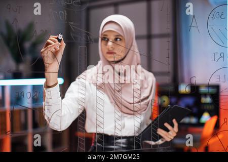 Focused smart muslim businesswoman, drawing graphs on glass presentation board. Female manager finance expert working in office at night, writing on glass board and using tablet computer Stock Photo