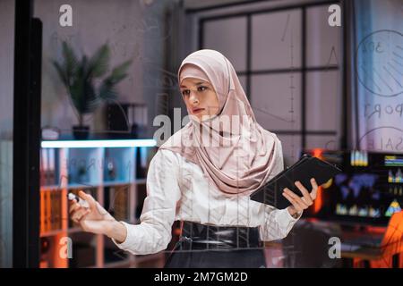 Focused smart muslim businesswoman, drawing graphs on glass presentation board. Female manager finance expert working in office at night, writing on glass board and using tablet computer Stock Photo