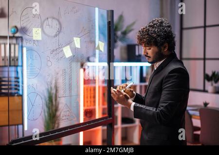 Focused male employee writing on colorful sticky notes on office glass board, developing business project, using tablet pc. Startup plan, people and technologies concept Stock Photo