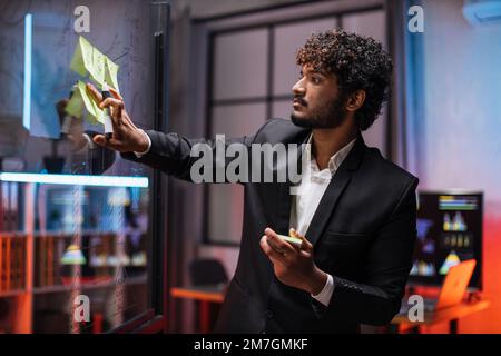 Focused male employee writing on colorful sticky notes on office glass board, developing business project, using tablet pc. Startup plan, people and technologies concept Stock Photo