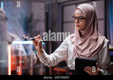 Side view of smart young businesswoman in hijab, brainstorming near office glass board, using tablet. Confident female employee writing on glass wall, developing business project at workplace. Stock Photo