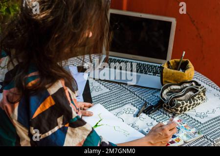 Freelance adult woman sitting at home designing a watercolor sketch for an online client, working with the laptop and drawing on white sheet. Stock Photo