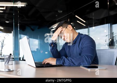 A tired young Asian man sits in the office at the table, holds his head, feels a headache, tension, migraine after a hard overtime day, shakes. Stock Photo