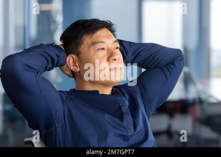 A calm young Asian man sits in the office on a chair, leans back, hands behind his head, looks thoughtfully and dreamily out the window. Resting from a hard day, satisfied with successful work. Stock Photo