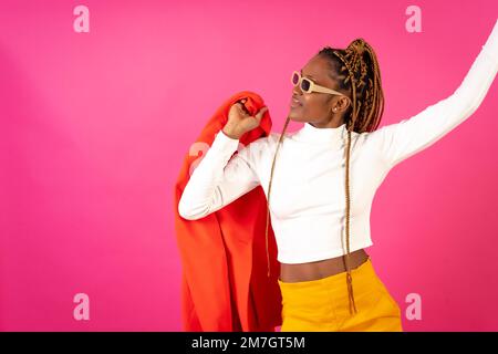 Black ethnic woman with braids on a pink background, wearing a red suit and white t-shirt dancing Stock Photo