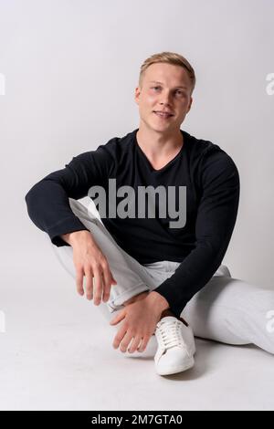 Caucasian blonde model in a black sweater on a white background sitting on the floor smiling Stock Photo