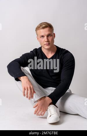 Caucasian blonde model in a black sweater on a white background, sitting on the floor Stock Photo
