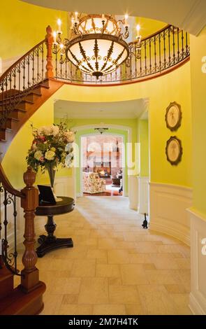 Curved wooden staircase and illuminated chandelier above hallway leading to living room with lit gas fireplace inside elegant cottage style home. Stock Photo