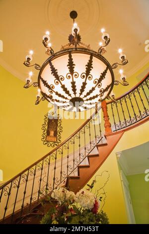 Illuminated chandelier with decorative details above hallway and curved wooden staircase inside lime green painted elegant cottage style home. Stock Photo