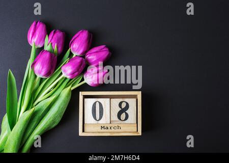 Bouquet of purple tulips and wooden block calendar with 8 march date on black table. Womens day holiday concept. Top view, flat lay, copy space. Stock Photo