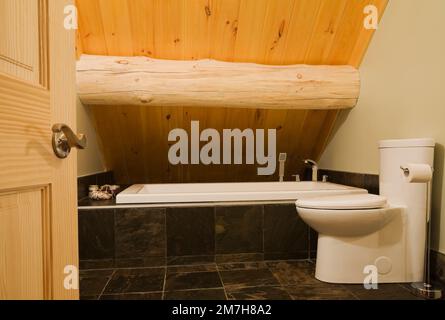 White porcelain toilet and ceramic encased bathtub in main bathroom on  upstairs floor inside luxurious Scandinavian style log home. Stock Photo