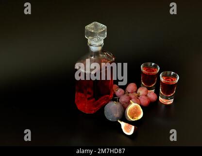 Fig-grape liquor, strong homemade alcohol in two glasses and a bottle on a black background, ripe fruits nearby. Close-up. Stock Photo