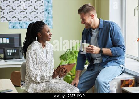 Side view portrait of two young colleagues chatting in office and smiling while taking break from work Stock Photo