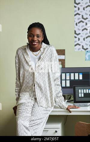 Vertical portrait of beautiful black woman smiling at camera while leaning on desk in home office Stock Photo