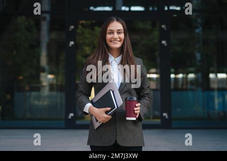 Happy brunette woman holding standing outside near office building with laptop, during coffee break. Smiling woman outdoor portrait. Modern lifestyle Stock Photo