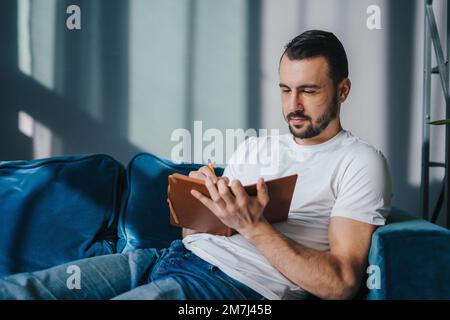 Young man sitting on couch writing down information in note book, taking important notes with pencil. Distance education. Planning and people concept Stock Photo
