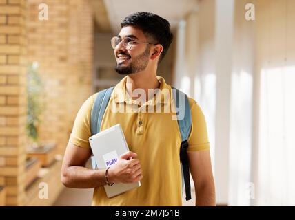 University student, college and indian man with a tablet and backpack while walking down campus corridor. Young gen z male happy about education Stock Photo