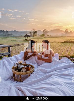 A couple of men and women at a cottage homestay in an outdoor bed in Northern Thailand Nan Province, couple waking up during sunrise in the rice fields in the morning drinking coffee outside Stock Photo