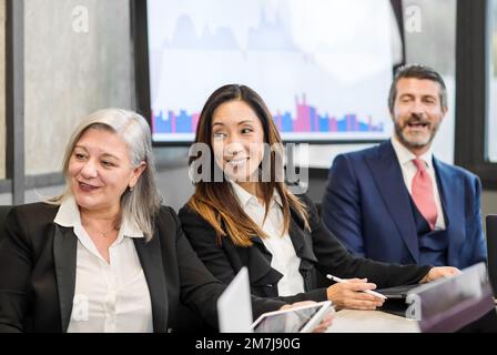 Multiracial colleagues in formal wear smiling and looking away while sitting at table during meeting in modern boardroom Stock Photo