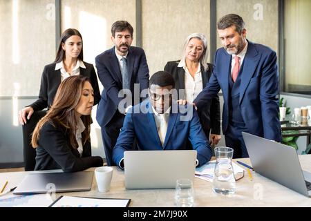 Serious black businessman in suit and glasses browsing data on netbook while sitting at table amidst multiracial colleagues in office Stock Photo