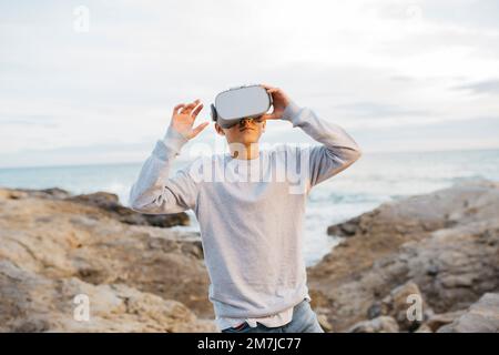 Boy Wearing Virtual Reality headset simulator on a rock breakwater, next to the sea Stock Photo