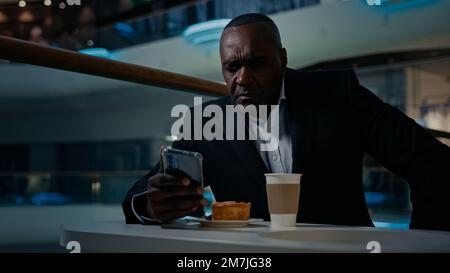 African American adult middle-aged ethnic man worker businessman employer entrepreneur eating drinking relax sitting at table in cafe drink coffee tea Stock Photo