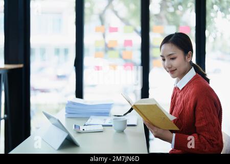 Portrait of a thoughtful Asian businesswoman looking at financial statements and making marketing plans using a computer with notebook on her desk Stock Photo